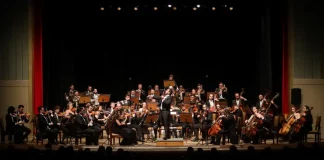 Músicos da Blumenau Filarmônica tocam em um palco com fundo escuro e cortina vermelha ao fundo. Eles estão concentrados em seus instrumentos, enquanto uma plateia de espectadores assiste à apresentação.
