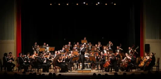Músicos da Blumenau Filarmônica tocam em um palco com fundo escuro e cortina vermelha ao fundo. Eles estão concentrados em seus instrumentos, enquanto uma plateia de espectadores assiste à apresentação.