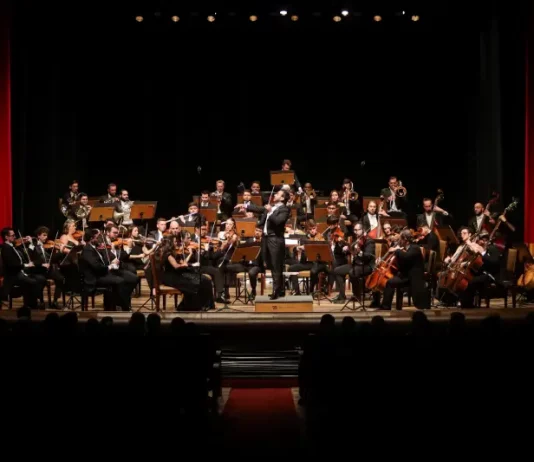 Músicos da Blumenau Filarmônica tocam em um palco com fundo escuro e cortina vermelha ao fundo. Eles estão concentrados em seus instrumentos, enquanto uma plateia de espectadores assiste à apresentação.