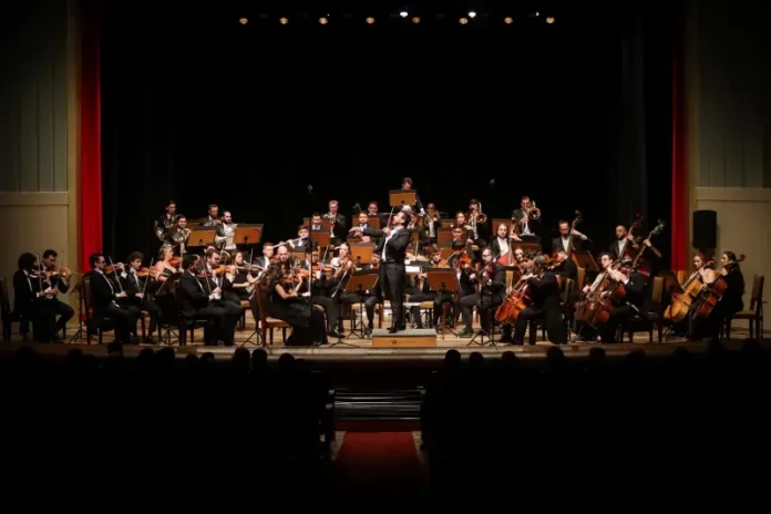 Músicos da Blumenau Filarmônica tocam em um palco com fundo escuro e cortina vermelha ao fundo. Eles estão concentrados em seus instrumentos, enquanto uma plateia de espectadores assiste à apresentação.