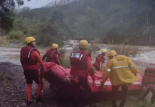 Equipe de cinco bombeiros com capacetes amarelos e roupas de proteção, carregando barco inflável rumo ao rio para resgate de três homens ilhados em Orleans