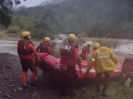 Equipe de cinco bombeiros com capacetes amarelos e roupas de proteção, carregando barco inflável rumo ao rio para resgate de três homens ilhados em Orleans