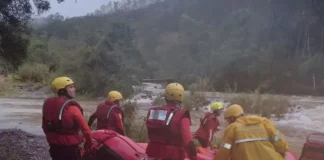 Equipe de cinco bombeiros com capacetes amarelos e roupas de proteção, carregando barco inflável rumo ao rio para resgate de três homens ilhados em Orleans