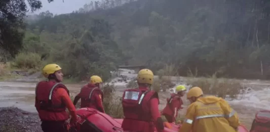 Equipe de cinco bombeiros com capacetes amarelos e roupas de proteção, carregando barco inflável rumo ao rio para resgate de três homens ilhados em Orleans