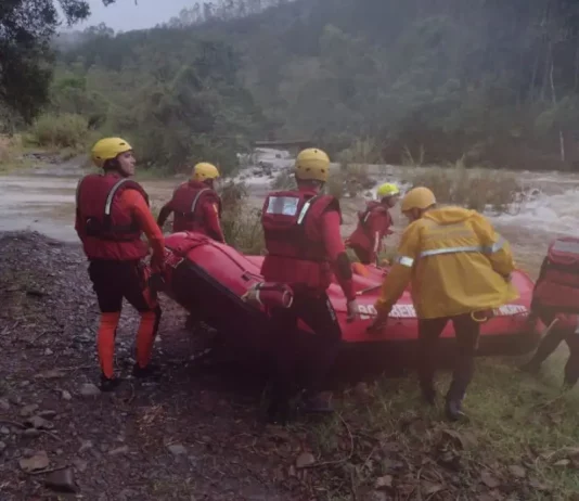 Equipe de cinco bombeiros com capacetes amarelos e roupas de proteção, carregando barco inflável rumo ao rio para resgate de três homens ilhados em Orleans