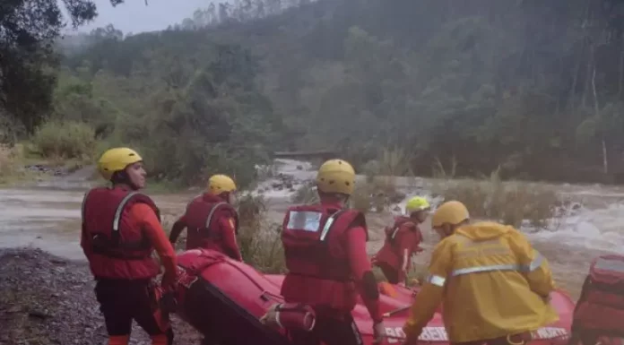 Equipe de cinco bombeiros com capacetes amarelos e roupas de proteção, carregando barco inflável rumo ao rio para resgate de três homens ilhados em Orleans