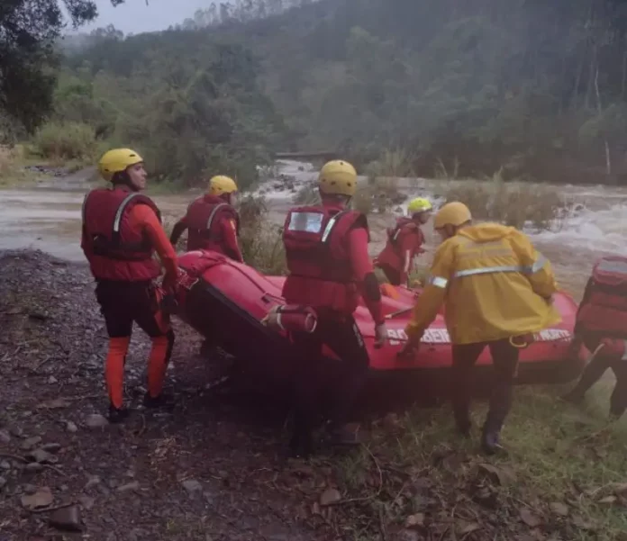 Equipe de cinco bombeiros com capacetes amarelos e roupas de proteção, carregando barco inflável rumo ao rio para resgate de três homens ilhados em Orleans