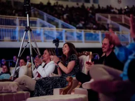 Plateia participa ativamente de uma apresentação na HJ Conference. Em primeiro plano, três pessoas estão em pé aplaudindo: uma mulher vestindo um vestido preto com contorno de flores brancas, uma mulher com casaco branco e calça cinza, e um homem com camisa preta. Ao lado da mulher de casaco branco, há um tripé com uma câmera de filmagem posicionada para registrar o evento. Ao fundo, várias pessoas estão sentadas, atentas à apresentação. O ambiente é fechado, iluminado, com estrutura típica de centro de eventos.