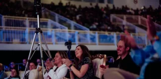 Plateia participa ativamente de uma apresentação na HJ Conference. Em primeiro plano, três pessoas estão em pé aplaudindo: uma mulher vestindo um vestido preto com contorno de flores brancas, uma mulher com casaco branco e calça cinza, e um homem com camisa preta. Ao lado da mulher de casaco branco, há um tripé com uma câmera de filmagem posicionada para registrar o evento. Ao fundo, várias pessoas estão sentadas, atentas à apresentação. O ambiente é fechado, iluminado, com estrutura típica de centro de eventos.