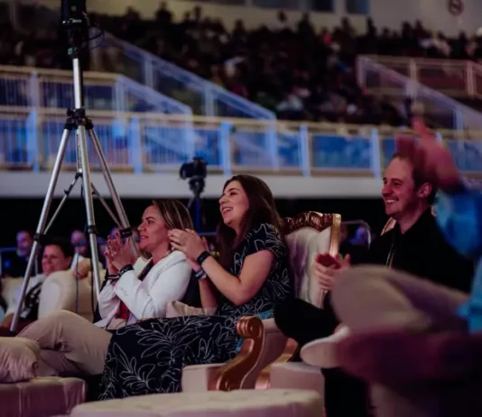 Plateia participa ativamente de uma apresentação na HJ Conference. Em primeiro plano, três pessoas estão em pé aplaudindo: uma mulher vestindo um vestido preto com contorno de flores brancas, uma mulher com casaco branco e calça cinza, e um homem com camisa preta. Ao lado da mulher de casaco branco, há um tripé com uma câmera de filmagem posicionada para registrar o evento. Ao fundo, várias pessoas estão sentadas, atentas à apresentação. O ambiente é fechado, iluminado, com estrutura típica de centro de eventos.