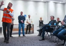 Michel Schlemper, vice-prefeito de São José, está sentado com camisa azul, blazer cinza e calça jeans, atento à apresentação. O secretário estadual de Infraestrutura, em pé, usa camisa branca, colete laranja com faixas cinzas e segura um microfone enquanto fala. Ao fundo, três homens e uma mulher estão em pé na lateral da sala, e dois homens sentados também prestam atenção