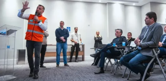 Michel Schlemper, vice-prefeito de São José, está sentado com camisa azul, blazer cinza e calça jeans, atento à apresentação. O secretário estadual de Infraestrutura, em pé, usa camisa branca, colete laranja com faixas cinzas e segura um microfone enquanto fala. Ao fundo, três homens e uma mulher estão em pé na lateral da sala, e dois homens sentados também prestam atenção
