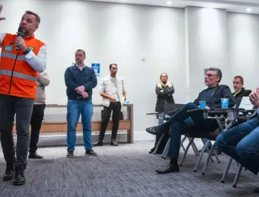 Michel Schlemper, vice-prefeito de São José, está sentado com camisa azul, blazer cinza e calça jeans, atento à apresentação. O secretário estadual de Infraestrutura, em pé, usa camisa branca, colete laranja com faixas cinzas e segura um microfone enquanto fala. Ao fundo, três homens e uma mulher estão em pé na lateral da sala, e dois homens sentados também prestam atenção