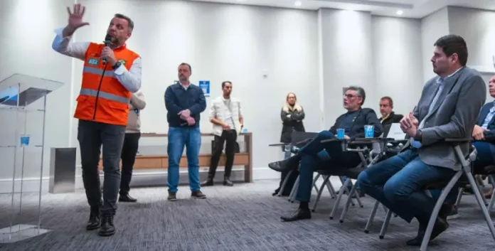 Michel Schlemper, vice-prefeito de São José, está sentado com camisa azul, blazer cinza e calça jeans, atento à apresentação. O secretário estadual de Infraestrutura, em pé, usa camisa branca, colete laranja com faixas cinzas e segura um microfone enquanto fala. Ao fundo, três homens e uma mulher estão em pé na lateral da sala, e dois homens sentados também prestam atenção