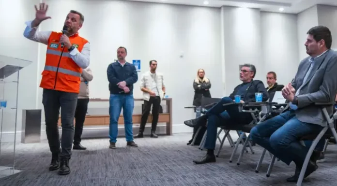 Michel Schlemper, vice-prefeito de São José, está sentado com camisa azul, blazer cinza e calça jeans, atento à apresentação. O secretário estadual de Infraestrutura, em pé, usa camisa branca, colete laranja com faixas cinzas e segura um microfone enquanto fala. Ao fundo, três homens e uma mulher estão em pé na lateral da sala, e dois homens sentados também prestam atenção