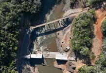 Foto aérea de uma usina hidrelétrica rodeada por árvores; o rio serpenteia pela paisagem e corre em direção à estrutura da usina, onde a força da água é usada para gerar energia elétrica.
