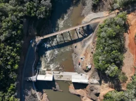 Foto aérea de uma usina hidrelétrica rodeada por árvores; o rio serpenteia pela paisagem e corre em direção à estrutura da usina, onde a força da água é usada para gerar energia elétrica.