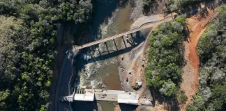 Foto aérea de uma usina hidrelétrica rodeada por árvores; o rio serpenteia pela paisagem e corre em direção à estrutura da usina, onde a força da água é usada para gerar energia elétrica.
