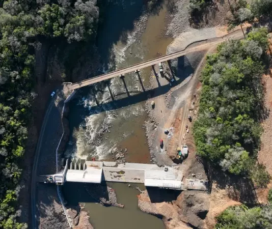 Foto aérea de uma usina hidrelétrica rodeada por árvores; o rio serpenteia pela paisagem e corre em direção à estrutura da usina, onde a força da água é usada para gerar energia elétrica.