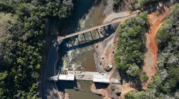 Foto aérea de uma usina hidrelétrica rodeada por árvores; o rio serpenteia pela paisagem e corre em direção à estrutura da usina, onde a força da água é usada para gerar energia elétrica.