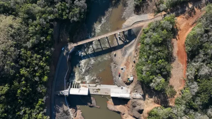 Foto aérea de uma usina hidrelétrica rodeada por árvores; o rio serpenteia pela paisagem e corre em direção à estrutura da usina, onde a força da água é usada para gerar energia elétrica.