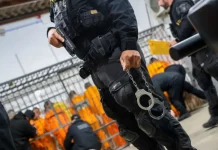 Escolta da Polícia Penal de SC: agente armado com pistola e algemas, com detentos ao fundo em operação de transferência.