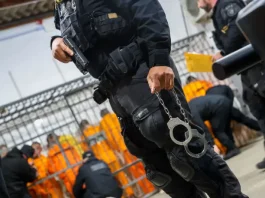 Escolta da Polícia Penal de SC: agente armado com pistola e algemas, com detentos ao fundo em operação de transferência.