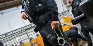 Escolta da Polícia Penal de SC: agente armado com pistola e algemas, com detentos ao fundo em operação de transferência.