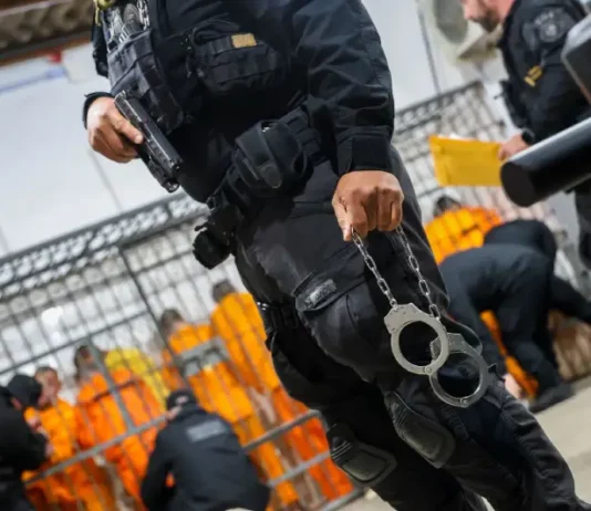 Escolta da Polícia Penal de SC: agente armado com pistola e algemas, com detentos ao fundo em operação de transferência.