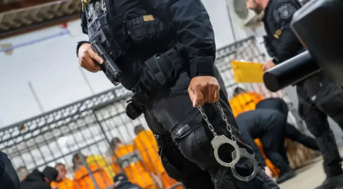 Escolta da Polícia Penal de SC: agente armado com pistola e algemas, com detentos ao fundo em operação de transferência.