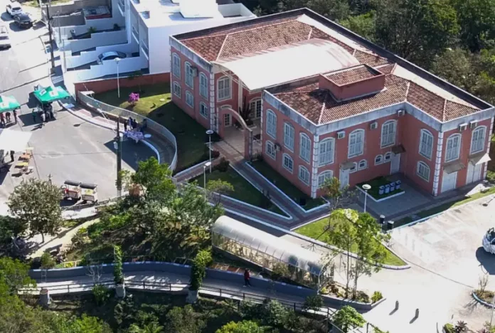Foto aérea da sede do Jardim Botânico de São José. A construção é de tijolos à vista, com telhado em duas águas. Em frente ao prédio, há árvores de médio porte que compõem a paisagem natural do local.