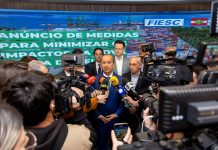 Na imagem, o governador Jorginho Mello está em pé, vestindo um terno escuro com camisa clara. Ele fala para a imprensa,. À sua volta, repórteres e assessores o cercam com microfones e gravações. Atrás dele, um banner azul exibe os logotipos da FIESC e do Governo de Santa Catarina