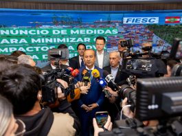 Na imagem, o governador Jorginho Mello está em pé, vestindo um terno escuro com camisa clara. Ele fala para a imprensa,. À sua volta, repórteres e assessores o cercam com microfones e gravações. Atrás dele, um banner azul exibe os logotipos da FIESC e do Governo de Santa Catarina