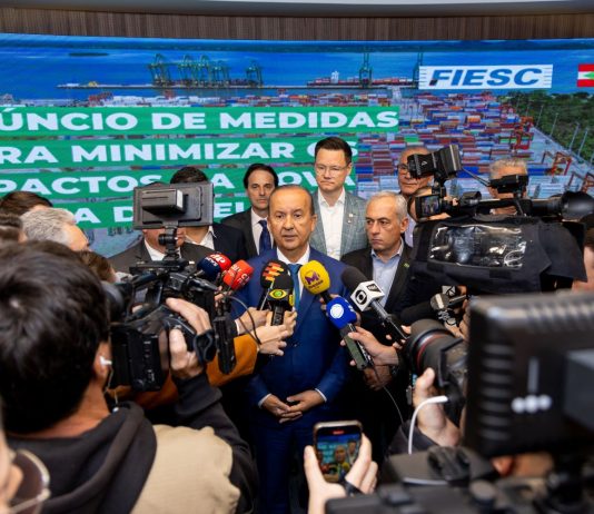 Na imagem, o governador Jorginho Mello está em pé, vestindo um terno escuro com camisa clara. Ele fala para a imprensa,. À sua volta, repórteres e assessores o cercam com microfones e gravações. Atrás dele, um banner azul exibe os logotipos da FIESC e do Governo de Santa Catarina