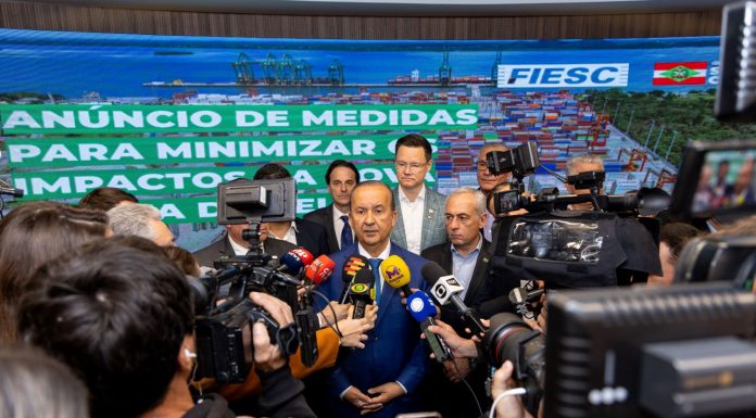 Na imagem, o governador Jorginho Mello está em pé, vestindo um terno escuro com camisa clara. Ele fala para a imprensa,. À sua volta, repórteres e assessores o cercam com microfones e gravações. Atrás dele, um banner azul exibe os logotipos da FIESC e do Governo de Santa Catarina