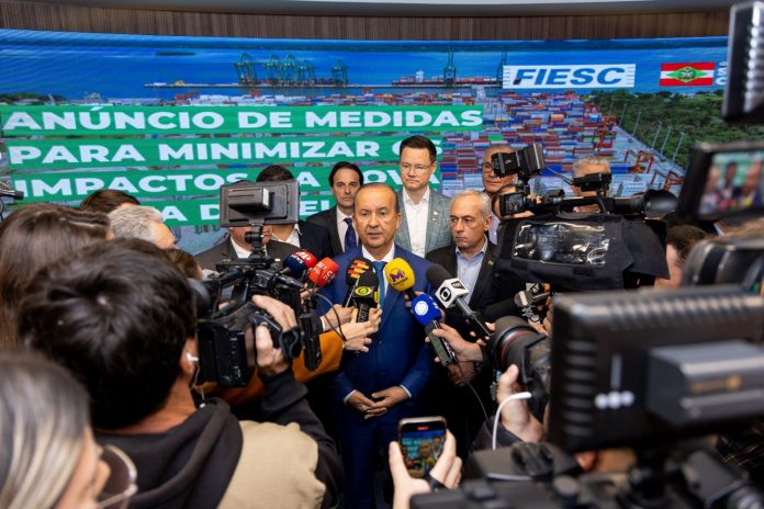 Na imagem, o governador Jorginho Mello está em pé, vestindo um terno escuro com camisa clara. Ele fala para a imprensa,. À sua volta, repórteres e assessores o cercam com microfones e gravações. Atrás dele, um banner azul exibe os logotipos da FIESC e do Governo de Santa Catarina