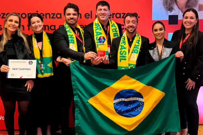 Representantes da Next Shipping posam segurando a bandeira do Brasil e exibindo medalhas e o certificado recebidos durante a cerimônia de premiação do ranking Great Place To Work 2025, em reconhecimento à excelência na gestão de pessoas.