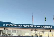 Na imagem, um carro preto da DEIC (Diretoria Estadual de Investigações Criminais), com faixas brancas nas laterais e o giroflex vermelho e azul ligado, está estacionado em frente ao prédio da Prefeitura de Palhoça. Dois policiais civis, vestidos com camisetas pretas com o emblema da Polícia Civil nas costas, descem do veículo. Ambos carregam documentos ou pastas nas mãos, aparentando estar em operação oficial. Ao fundo, é possível ver parte da fachada do prédio da prefeitura. A cena transmite um clima de ação policial e investigação.