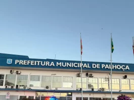 Na imagem, um carro preto da DEIC (Diretoria Estadual de Investigações Criminais), com faixas brancas nas laterais e o giroflex vermelho e azul ligado, está estacionado em frente ao prédio da Prefeitura de Palhoça. Dois policiais civis, vestidos com camisetas pretas com o emblema da Polícia Civil nas costas, descem do veículo. Ambos carregam documentos ou pastas nas mãos, aparentando estar em operação oficial. Ao fundo, é possível ver parte da fachada do prédio da prefeitura. A cena transmite um clima de ação policial e investigação.
