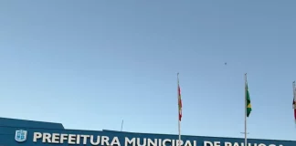 Na imagem, um carro preto da DEIC (Diretoria Estadual de Investigações Criminais), com faixas brancas nas laterais e o giroflex vermelho e azul ligado, está estacionado em frente ao prédio da Prefeitura de Palhoça. Dois policiais civis, vestidos com camisetas pretas com o emblema da Polícia Civil nas costas, descem do veículo. Ambos carregam documentos ou pastas nas mãos, aparentando estar em operação oficial. Ao fundo, é possível ver parte da fachada do prédio da prefeitura. A cena transmite um clima de ação policial e investigação.