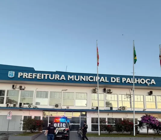 Na imagem, um carro preto da DEIC (Diretoria Estadual de Investigações Criminais), com faixas brancas nas laterais e o giroflex vermelho e azul ligado, está estacionado em frente ao prédio da Prefeitura de Palhoça. Dois policiais civis, vestidos com camisetas pretas com o emblema da Polícia Civil nas costas, descem do veículo. Ambos carregam documentos ou pastas nas mãos, aparentando estar em operação oficial. Ao fundo, é possível ver parte da fachada do prédio da prefeitura. A cena transmite um clima de ação policial e investigação.