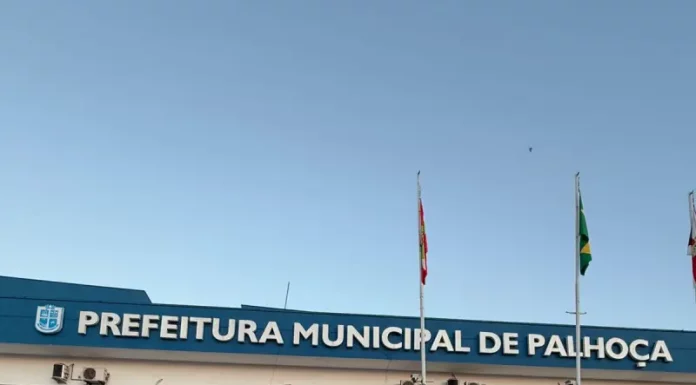 Na imagem, um carro preto da DEIC (Diretoria Estadual de Investigações Criminais), com faixas brancas nas laterais e o giroflex vermelho e azul ligado, está estacionado em frente ao prédio da Prefeitura de Palhoça. Dois policiais civis, vestidos com camisetas pretas com o emblema da Polícia Civil nas costas, descem do veículo. Ambos carregam documentos ou pastas nas mãos, aparentando estar em operação oficial. Ao fundo, é possível ver parte da fachada do prédio da prefeitura. A cena transmite um clima de ação policial e investigação.