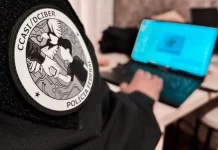 Homem sentado trabalha em um notebook. Em primeiro plano, aparece seu braço direito com uma jaqueta preta. No braço, há um logotipo redondo com a inscrição ‘CCASI/DEIBER’ e, logo abaixo, as palavras ‘Polícia Federal