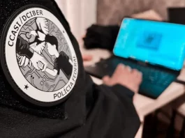 Homem sentado trabalha em um notebook. Em primeiro plano, aparece seu braço direito com uma jaqueta preta. No braço, há um logotipo redondo com a inscrição ‘CCASI/DEIBER’ e, logo abaixo, as palavras ‘Polícia Federal