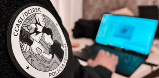 Homem sentado trabalha em um notebook. Em primeiro plano, aparece seu braço direito com uma jaqueta preta. No braço, há um logotipo redondo com a inscrição ‘CCASI/DEIBER’ e, logo abaixo, as palavras ‘Polícia Federal