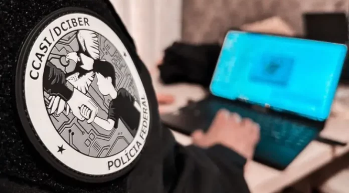 Homem sentado trabalha em um notebook. Em primeiro plano, aparece seu braço direito com uma jaqueta preta. No braço, há um logotipo redondo com a inscrição ‘CCASI/DEIBER’ e, logo abaixo, as palavras ‘Polícia Federal