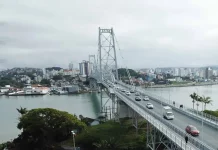 Foto aérea da Ponte Hercílio Luz em Florianópolis. A ponte, que liga o continente à Ilha, aparece com diversos veículos em circulação e algumas pessoas caminhando. Ao fundo, é possível ver o bairro Estreito, com vários prédios e construções residenciais. O mar contorna a estrutura da ponte e completa a paisagem urbana.
