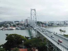 Foto aérea da Ponte Hercílio Luz em Florianópolis. A ponte, que liga o continente à Ilha, aparece com diversos veículos em circulação e algumas pessoas caminhando. Ao fundo, é possível ver o bairro Estreito, com vários prédios e construções residenciais. O mar contorna a estrutura da ponte e completa a paisagem urbana.