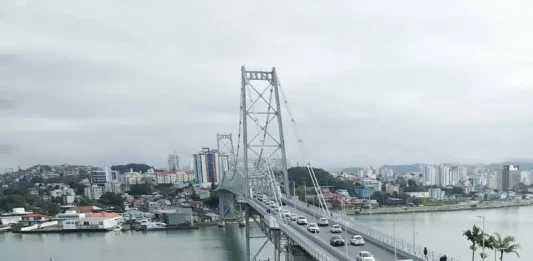 Foto aérea da Ponte Hercílio Luz em Florianópolis. A ponte, que liga o continente à Ilha, aparece com diversos veículos em circulação e algumas pessoas caminhando. Ao fundo, é possível ver o bairro Estreito, com vários prédios e construções residenciais. O mar contorna a estrutura da ponte e completa a paisagem urbana.