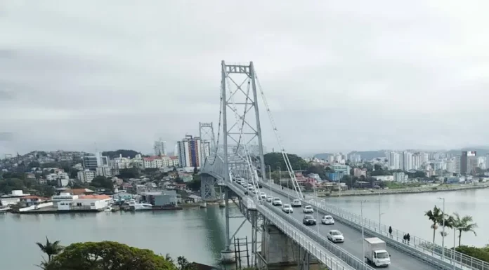 Foto aérea da Ponte Hercílio Luz em Florianópolis. A ponte, que liga o continente à Ilha, aparece com diversos veículos em circulação e algumas pessoas caminhando. Ao fundo, é possível ver o bairro Estreito, com vários prédios e construções residenciais. O mar contorna a estrutura da ponte e completa a paisagem urbana.
